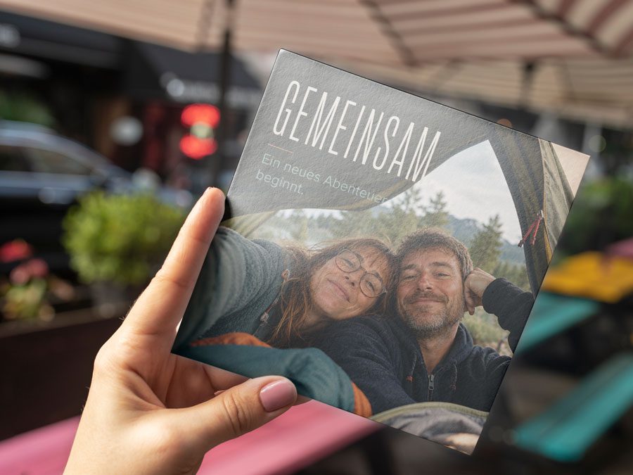 Hand hält die aufgeklappte Hochzeitseinladung „GEMEINSAM“ mit natürlichem Paarfoto im Zelt und dem Schriftzug „Ein neues Abenteuer beginnt.“